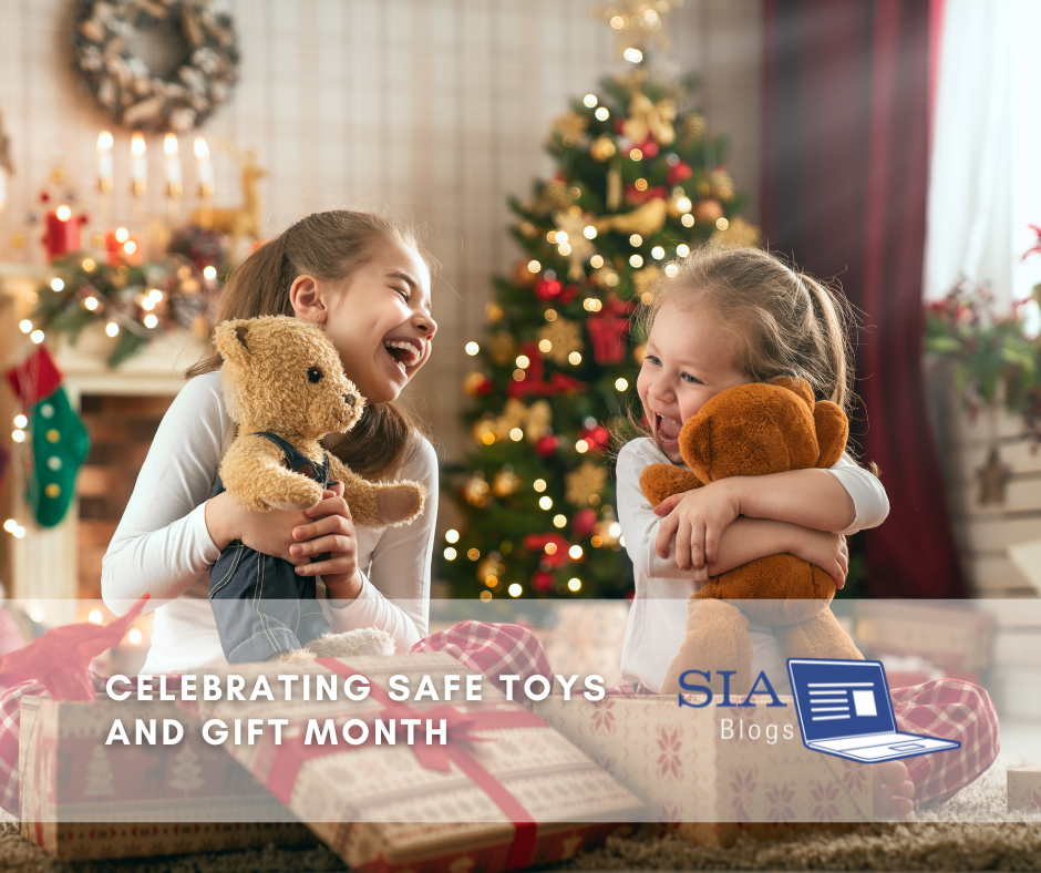 Two young girls joyfully hug teddy bears in front of a beautifully decorated Christmas tree, with gifts and festive decorations surrounding them in a cozy holiday setting. The caption reads, "Celebrating Safe Toys and Gift Month," along with the SIA Blogs logo.