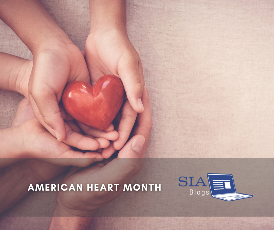 Several hands gently hold a red heart-shaped object, symbolizing love and care. The image conveys a message of compassion, particularly in the context of American Heart Month.