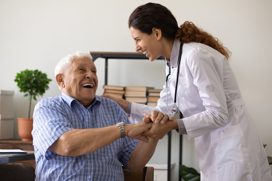 A cheerful doctor and an elderly patient share a warm moment in a medical office. The doctor, dressed in a white coat with a stethoscope, is laughing and holding the patient's hands, who is smiling broadly in response, creating a friendly and supportive atmosphere.
