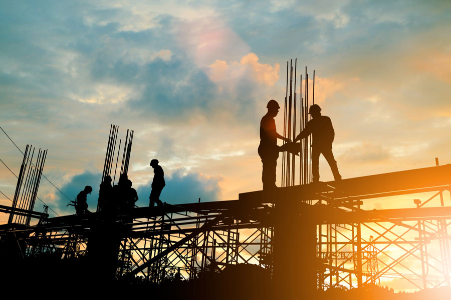 Silhouettes of construction workers collaborating on a building site against a vibrant sunset. The workers are engaged in tasks on scaffolding, with rebar protruding and a colorful sky in the background, highlighting the end of the workday.
