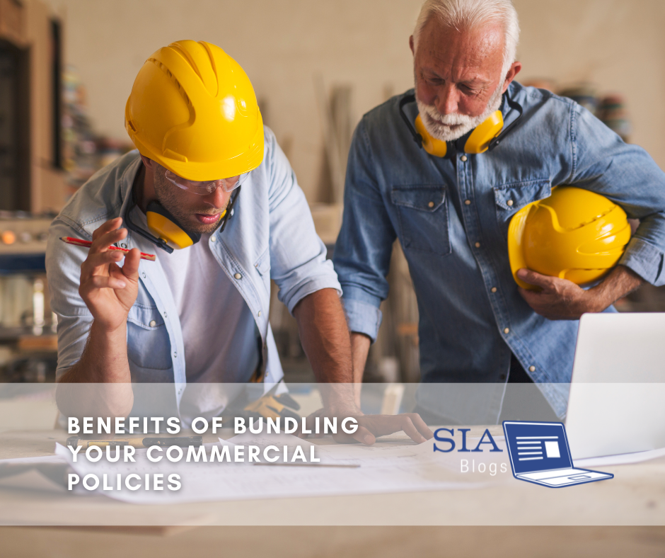 Two construction workers, both wearing yellow hard hats and safety gear, review plans on a table. One is pointing at the documents, while the other holds a hard hat and observes. The caption reads, "Benefits of Bundling Your Commercial Policies," along with the SIA Blogs logo.