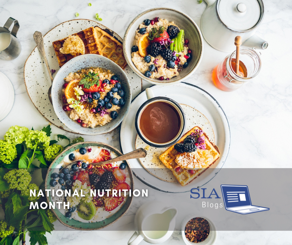 A vibrant breakfast spread featuring bowls of oatmeal topped with fresh fruits, waffles drizzled with syrup, and a cup of coffee arranged on a white table. The caption reads, "National Nutrition Month," with the SIA Blogs logo.