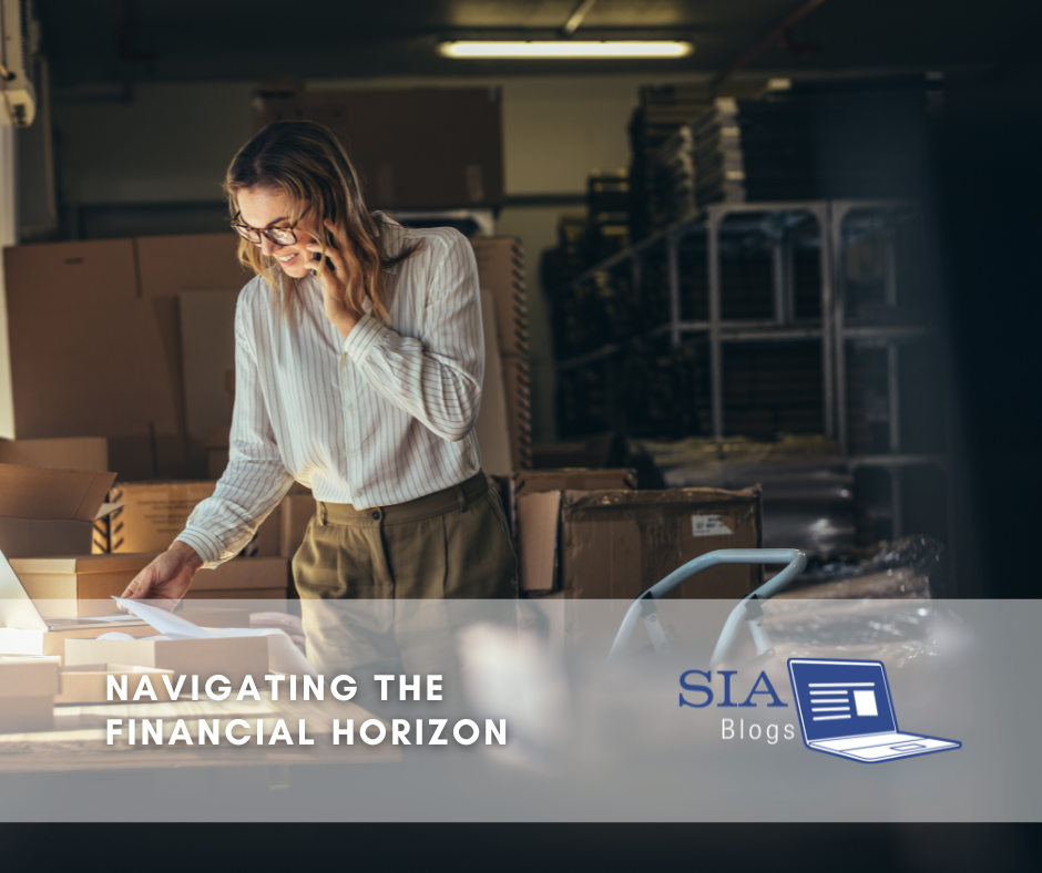A woman in a striped blouse is engaged in a phone call while reviewing paperwork in a warehouse filled with stacked boxes. Her expression suggests she is focused and organized, reflecting a proactive approach to managing her business.