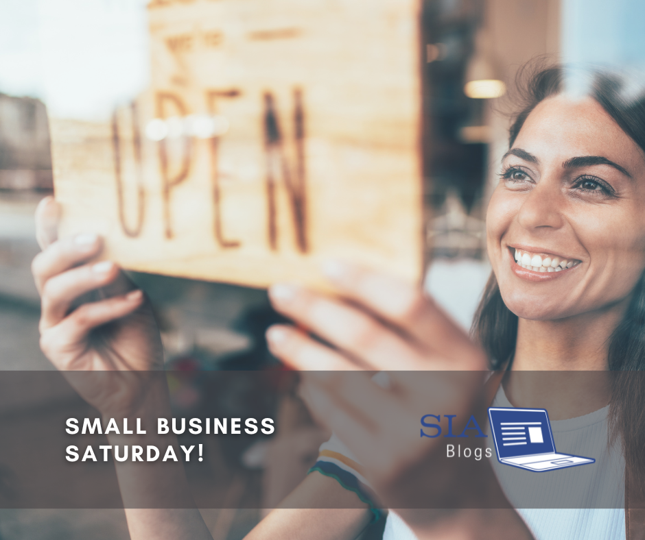 A smiling woman stands in front of her shop, holding up a wooden "Open" sign with enthusiasm. Her cheerful expression reflects the excitement of welcoming customers, emphasizing the spirit of Small Business Saturday.