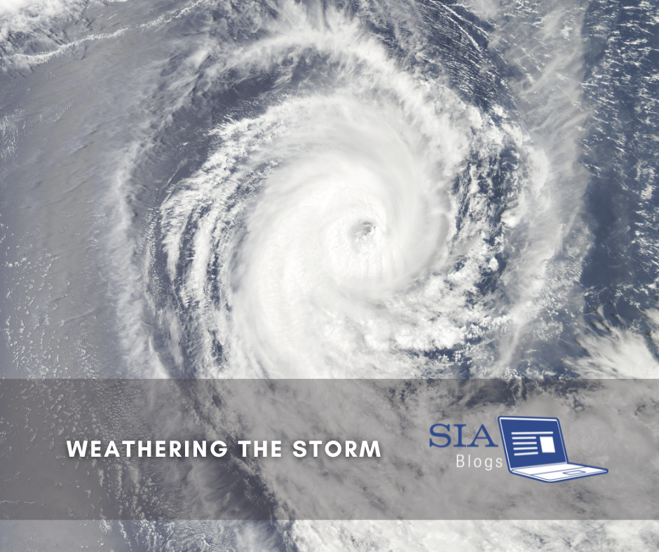 A swirling hurricane is captured from above, showing the intense spiral of clouds in the storm's eye as it moves across the ocean. The caption reads, "Weathering the Storm," with the SIA Blogs logo.